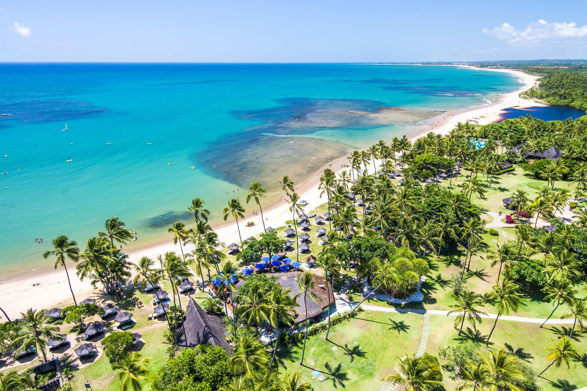 Aerial view of a tropical beach in Brazil with turquoise water, palm trees, and a resort with thatched roofs—perfect for a relaxing getaway.