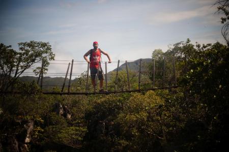 A tourist stands on a narrow suspension bridge in Chapada dos Veadeiros, surrounded by tropical nature.